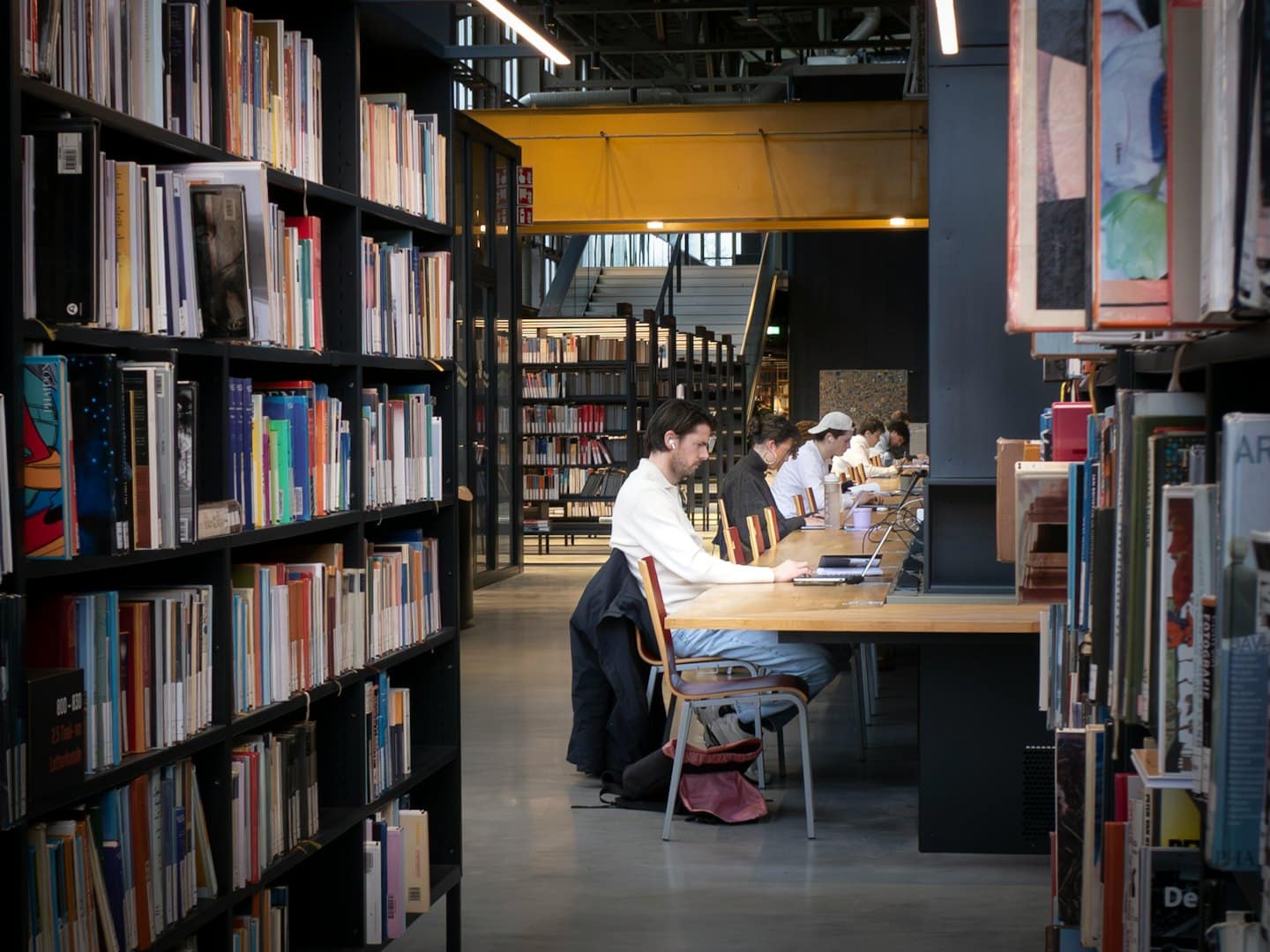 Students study on laptops at a long wooden library table.