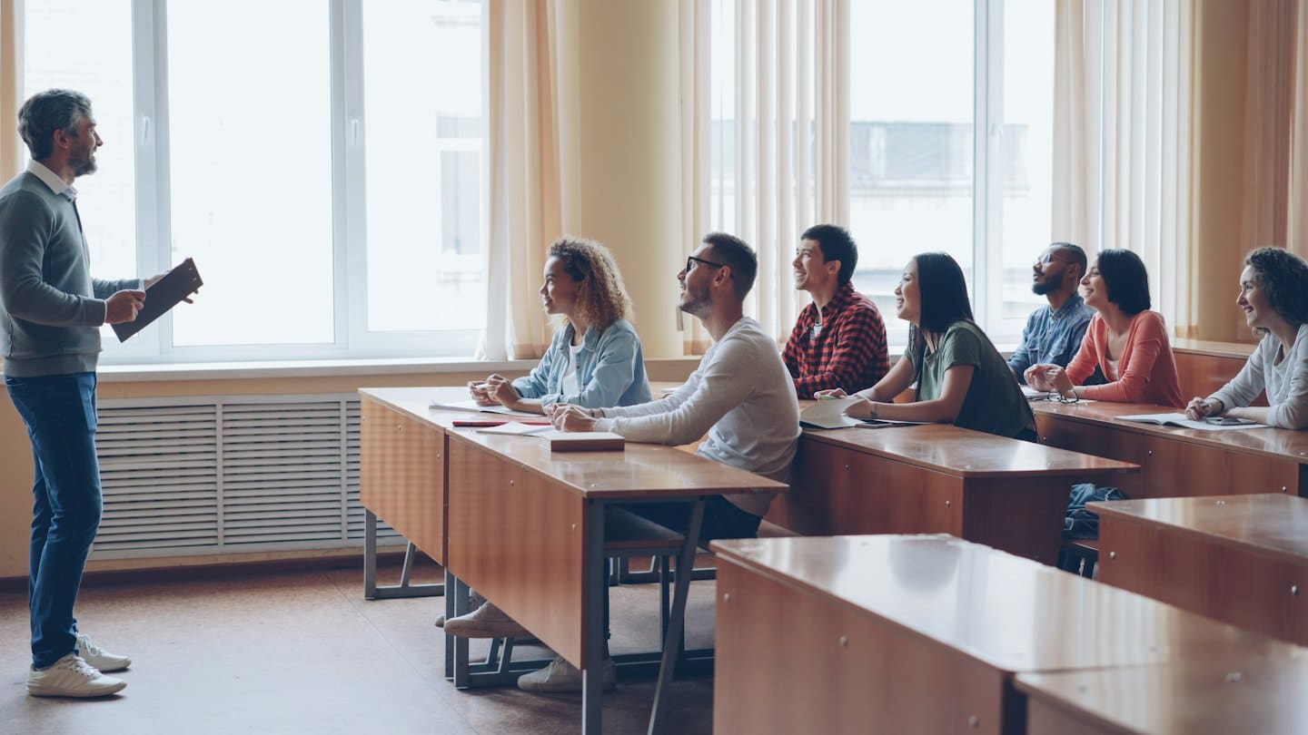 Instructor with clipboard lectures to attentive college students in a classroom.