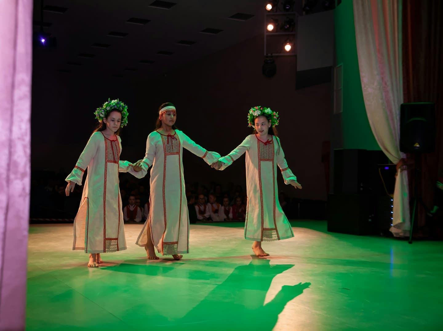 Three girls in embroidered dresses dance holding hands on stage.